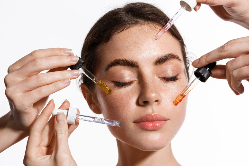 Porträt einer Frau, an deren Gesicht mehrere Pipetten mit pflegenden Seren und Hyaluron angesetzt werden – Wirkstoffkosmetik bei Donatella Beauty in Berlin Grunewald